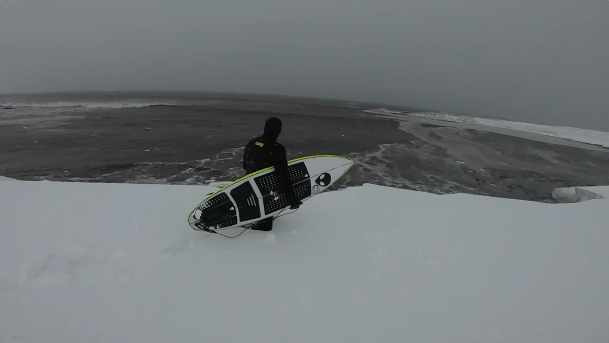 Surfer walking across snow into the Arctic ocean with a Lunasurf board — cold water surfing in Iceland